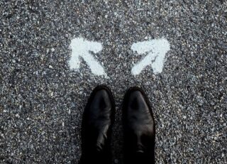 Looking down at dress shoes on pavement marked with two arrows at 90 degree angle, indicating choice.