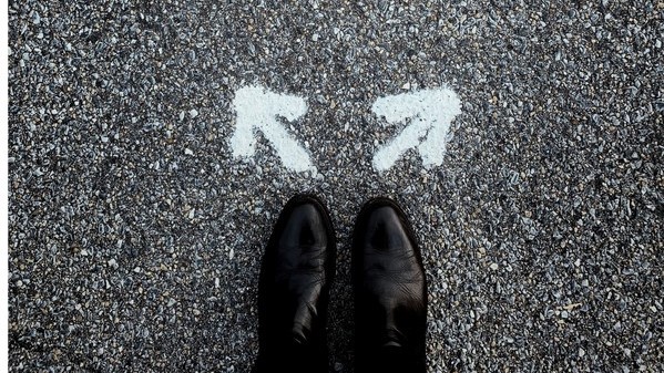 Looking down at dress shoes on pavement marked with two arrows at 90 degree angle, indicating choice.