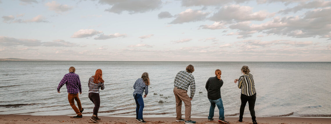 Six people on a sandy beach facing a lake, in motion skipping stones.