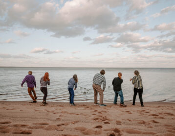 Six people on a sandy beach facing a lake, in motion skipping stones.